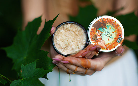 Hands holding an open jar of Barefoot Venus Maple Blondie sugar scrub, with syrup dripping from the fingers. The jar lid features a fall-themed design with a pin-up illustration, maple leaves, and a moose.